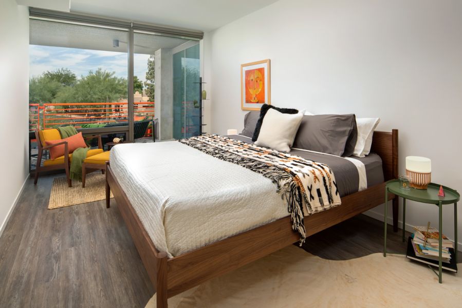 Modern bedroom with a wooden bed, balcony view, armchair, and decorative touches like a patterned blanket at Optima Sonoran Village apartments.