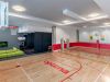 Indoor gym with a basketball hoop, mini tennis net, red markings, and a green couch near a TV in the corner.