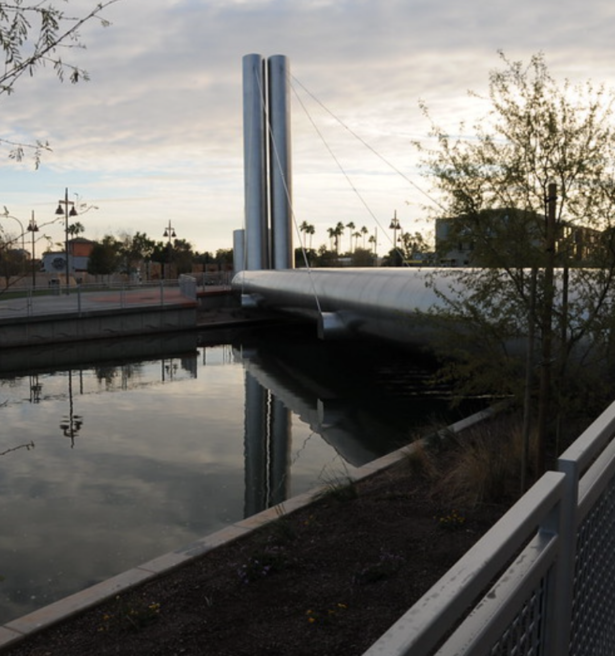Modern Soleri Bridge with tall silver pillars spans a canal in Scottsdale, surrounded by plants and glowing at sunset.