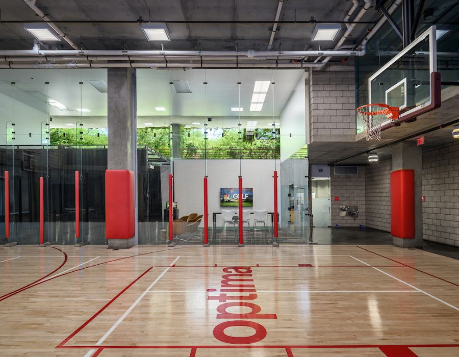 Indoor basketball court with red accents, glass walls, and a modern lounge area in the background.