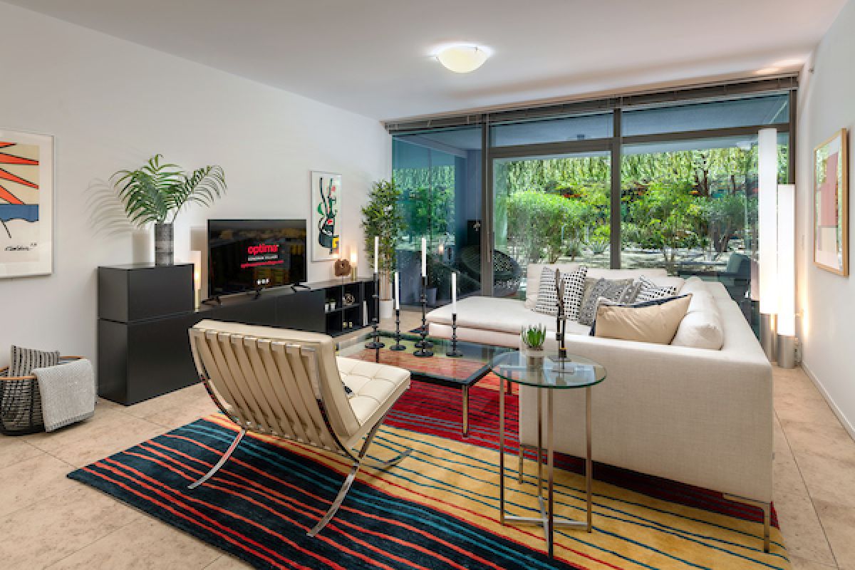 Modern living room at Optima Sonoran Village with white sectional sofa, striped rug, glass wall, and greenery outside.