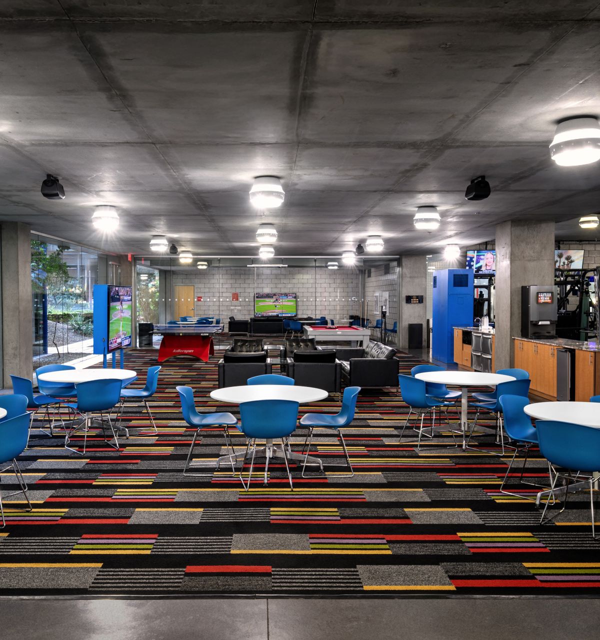 Modern lounge with blue chairs, round tables, colorful carpet, vending machines, and large windows overlooking greenery.