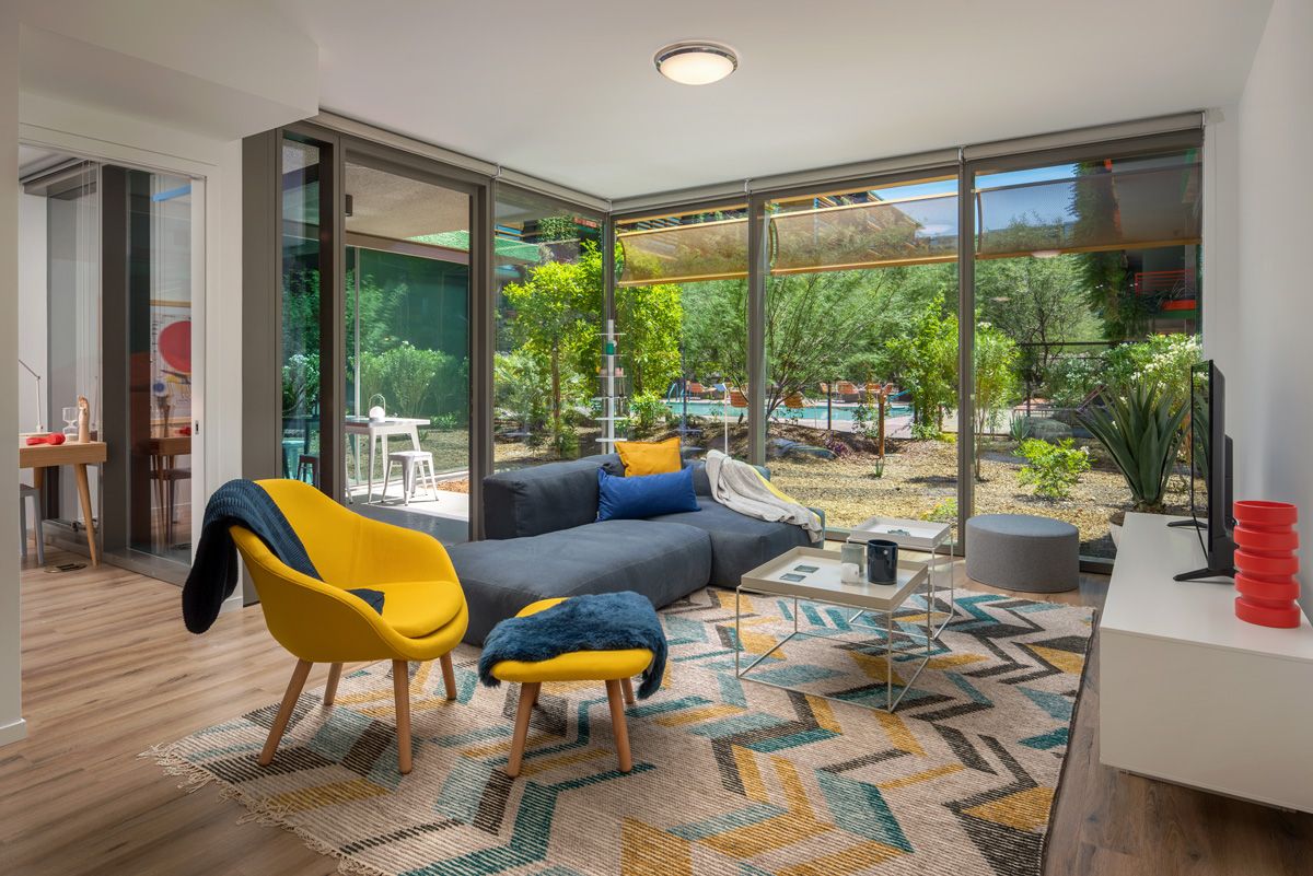 Optima Sonoran Village apartment Modern living room with large windows, yellow chairs, gray sofa, geometric rug, and view of a lush garden.