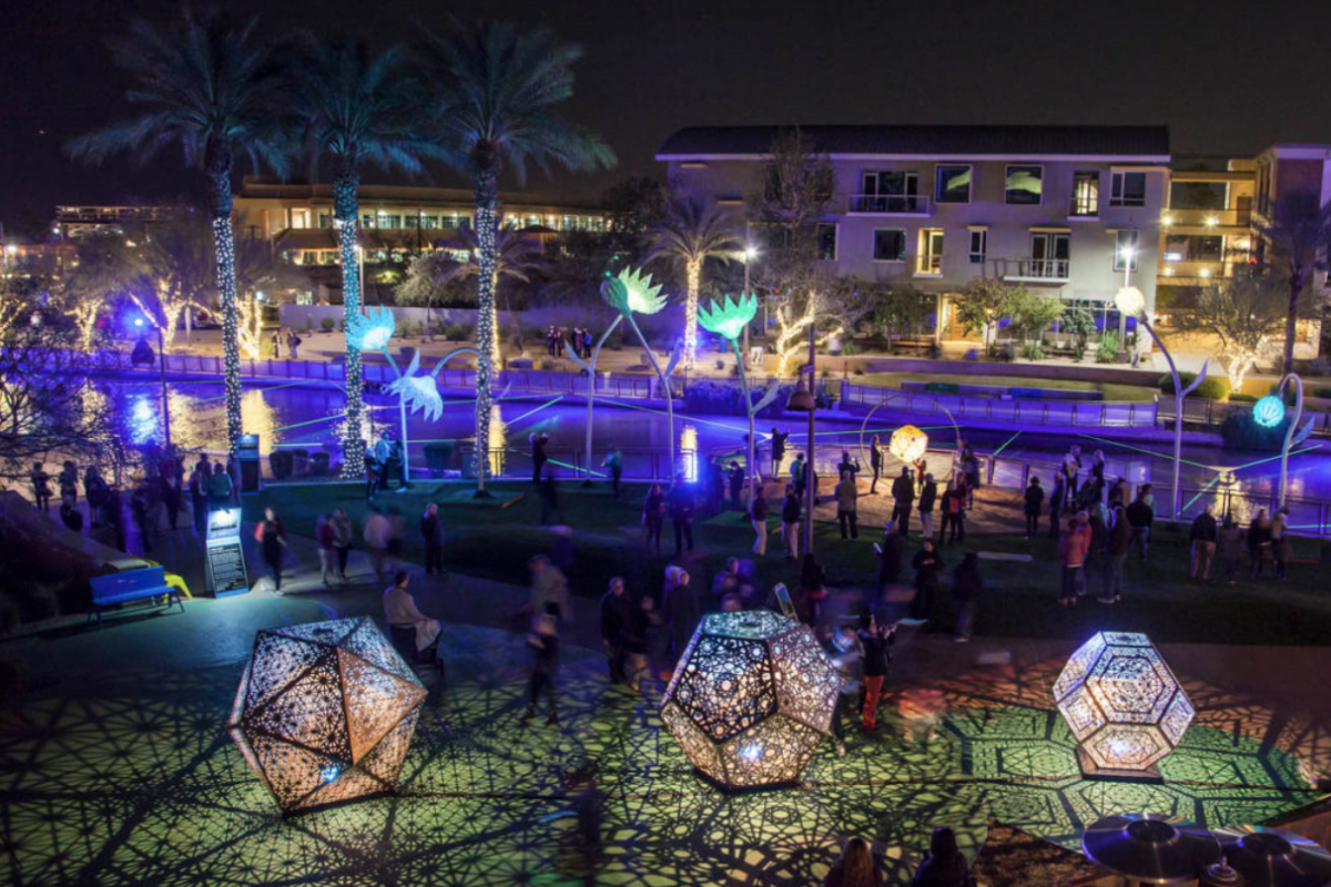 People walk among glowing geometric sculptures and palm trees at the 2023 Canal Convergence nighttime art festival near a canal.