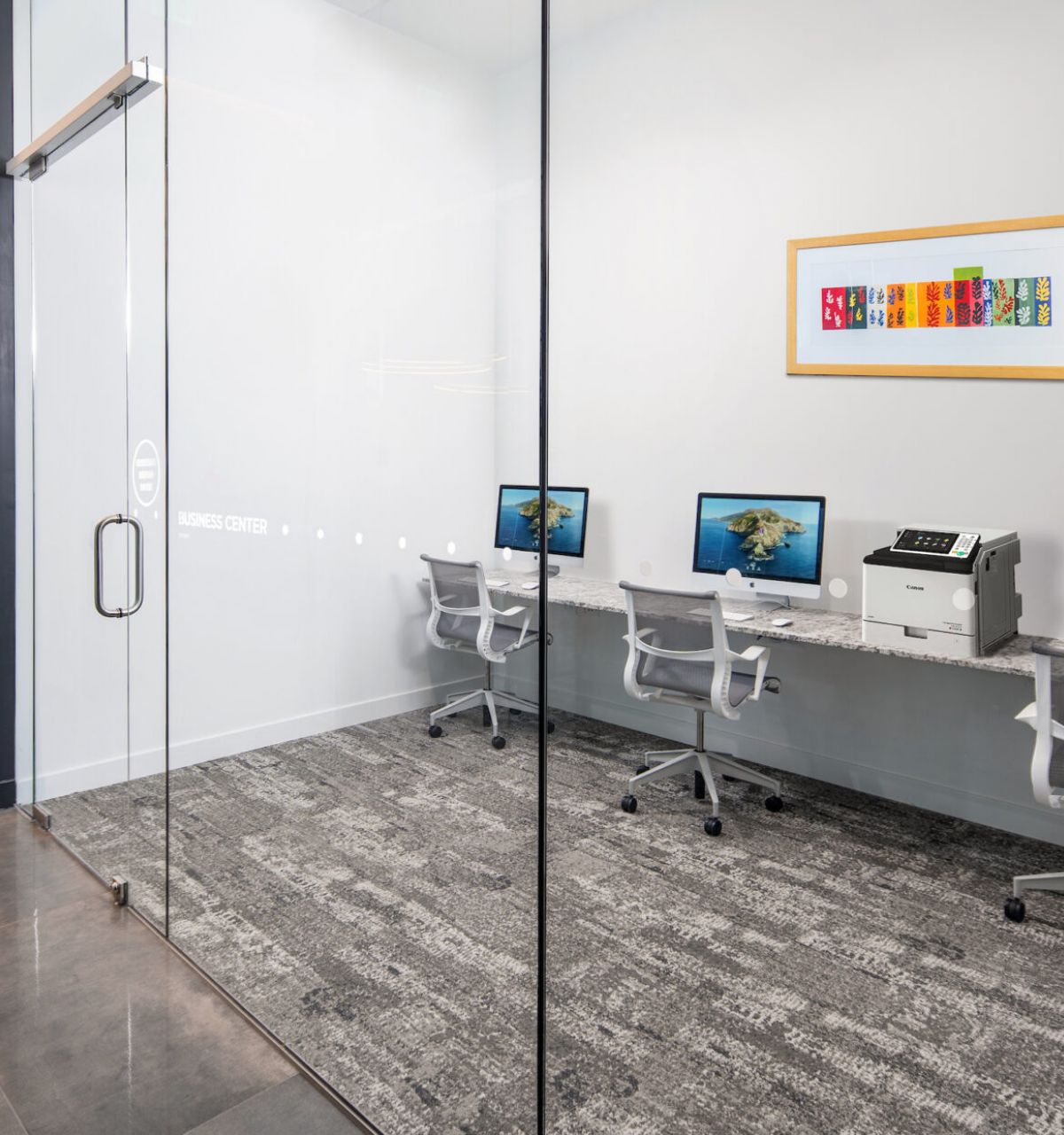 A modern business center by Optima Communities with computers, chairs, printer, and artwork—ideal for hybrid working behind glass doors.