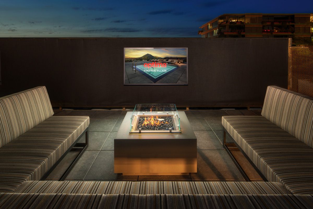 Rooftop patio with striped couches, a central fire pit, and a large screen at Optima Sonoran Village apartments.