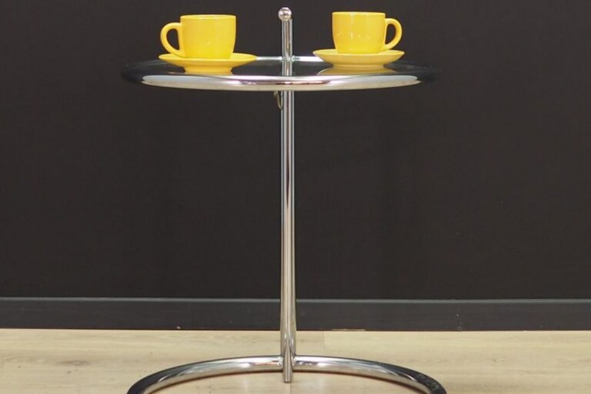 A spotlight shines on a round chrome Table E-1027 with two yellow cups, set against a black wall and wooden floor.
