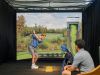 Two people enjoy daily life at Optima Community, using an indoor golf simulator—one swings while the other watches by the golf bag.