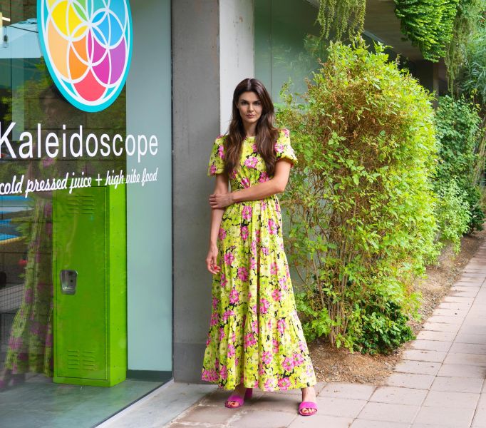 Woman in a floral dress stands by a glass door labeled "Kaleidoscope;" greenery surrounds the entrance at Optima Sonoran Village apartments.