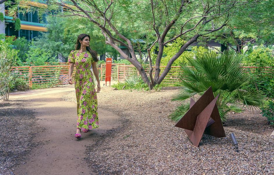 Woman in floral dress walks along a garden path with trees and a metal sculpture nearby at Optima Sonoran Village apartments.