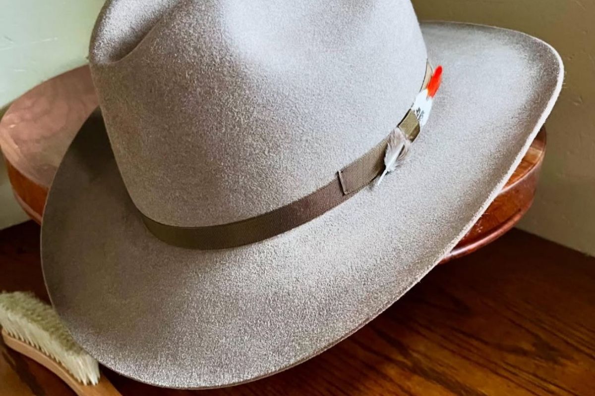 A gray fedora hat with a feather, perfect for Scottsdale hat bars, sits on a wooden table next to a hat brush.