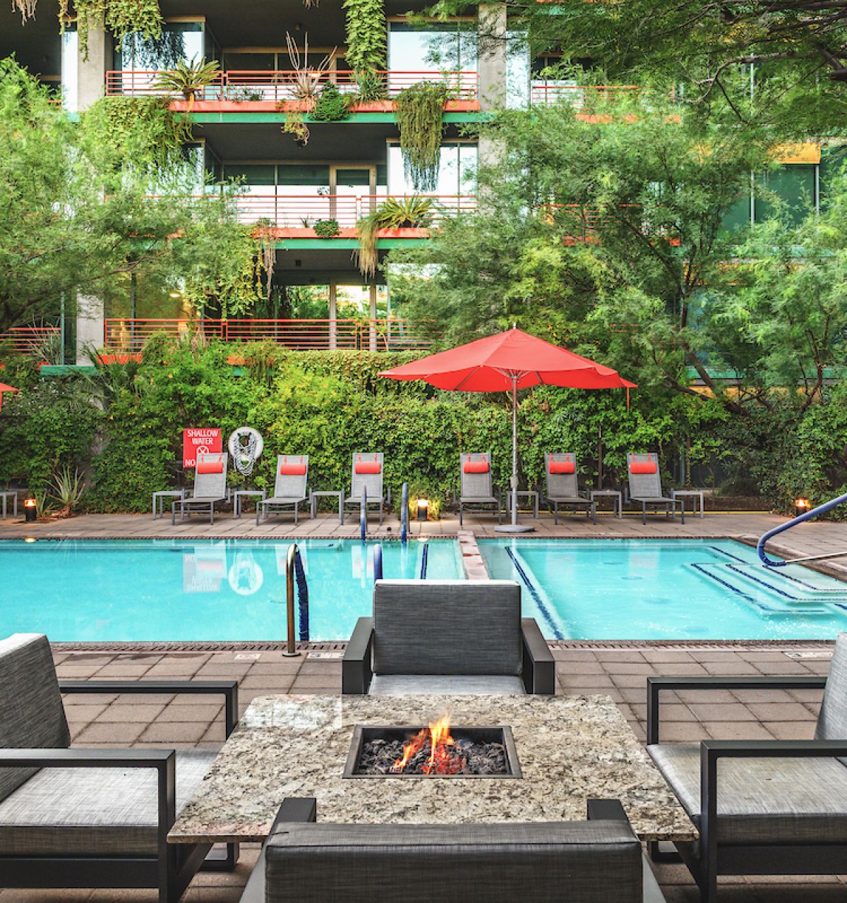 Modern poolside patio with red umbrellas, lounge chairs, and fire pit—perfect for Sky Deck evenings amid lush greenery in the background.