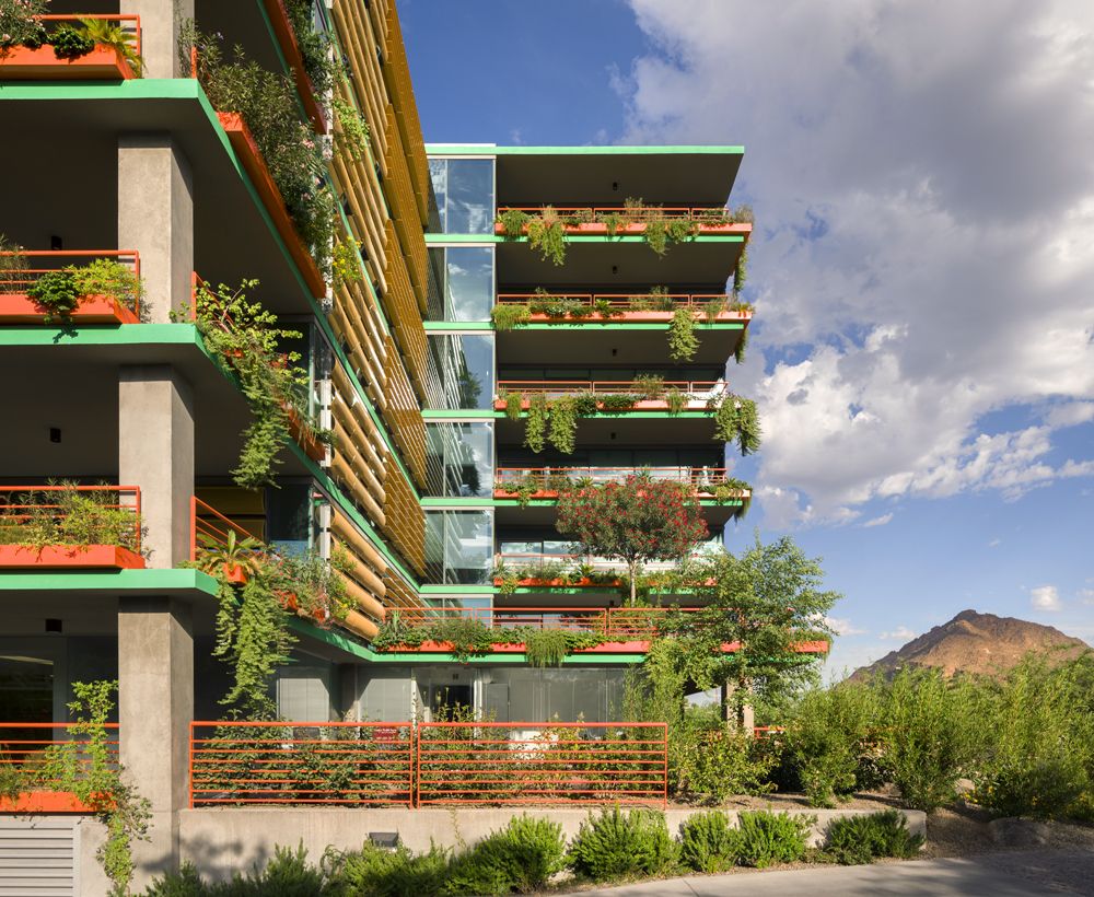 Modern Optima Sonoran Village apartment building featuring tiered balconies with orange railings, vertical slats, and abundant greenery.