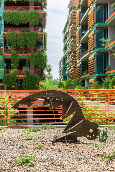 Artistic metal sculpture resembling an animal, with green buildings and orange railings in the background at Optima Sonoran Village apartments.