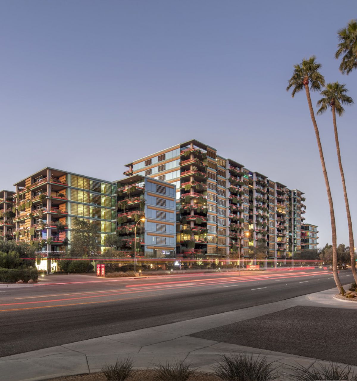 Modern multi-story apartment buildings at dusk, with palm trees and light trails from passing cars in the foreground.