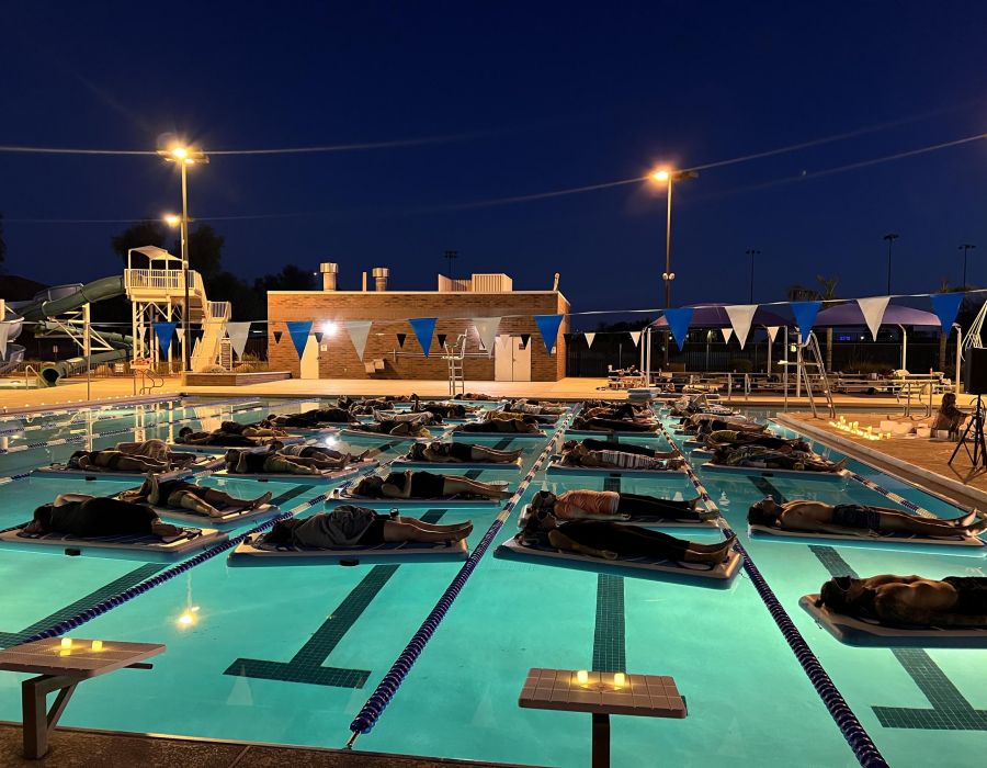 People lying on inflatable mats in a lit swimming pool at night, under a dark sky with poolside lights at Optima Sonoran Village apartments.