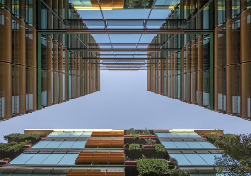 Looking up at Optima Sonoran Village apartments; a symmetrical, modern building with glass, greenery, and wooden elements under a clear sky.