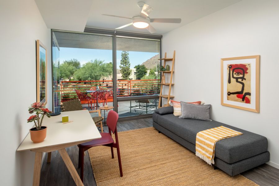 Minimalist room with a desk, red chair, gray sofa, and artwork near a large window showing an outdoor balcony and trees at Optima Sonoran Village apartments.