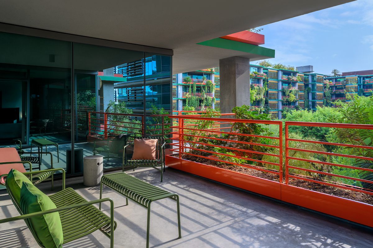 Modern Optima balcony with green metal furniture, orange railing, lush greenery, and buildings in the background enhances your outdoor space.