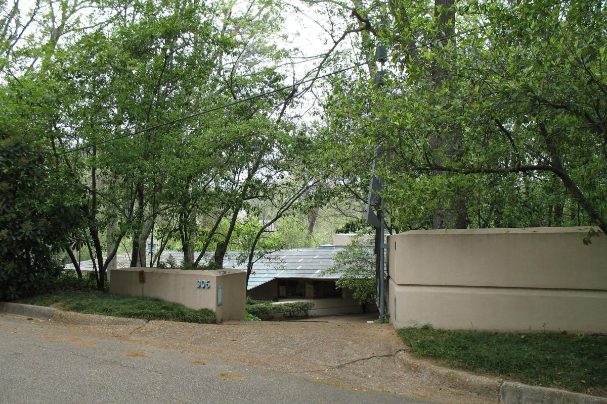 A modernism-inspired house with a curved beige wall and trees partially obscuring the front, viewed from the street.