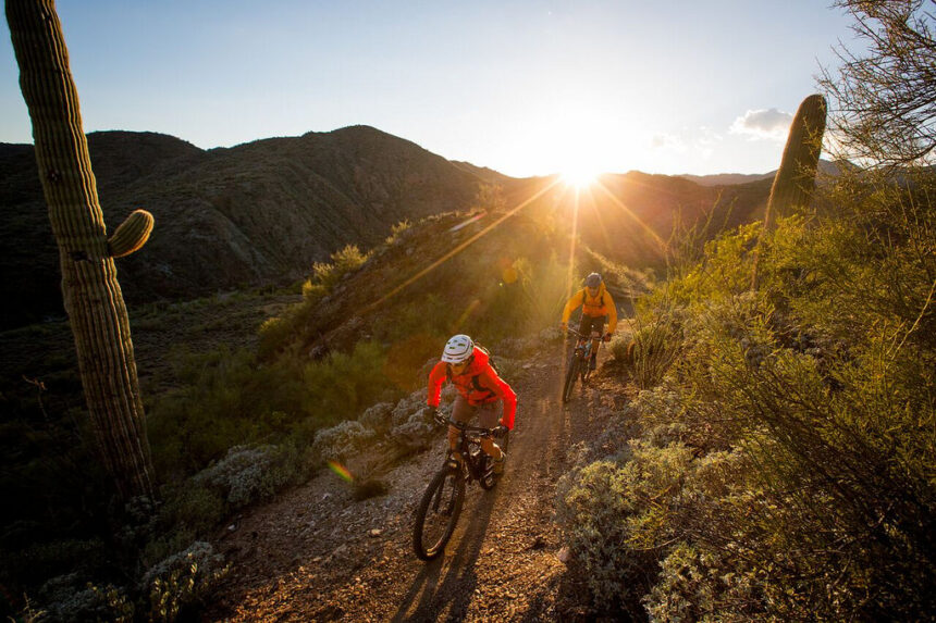 Two cyclists enjoy low-carbon, sustainable travel on a desert trail at sunset, surrounded by cacti and rugged mountains.