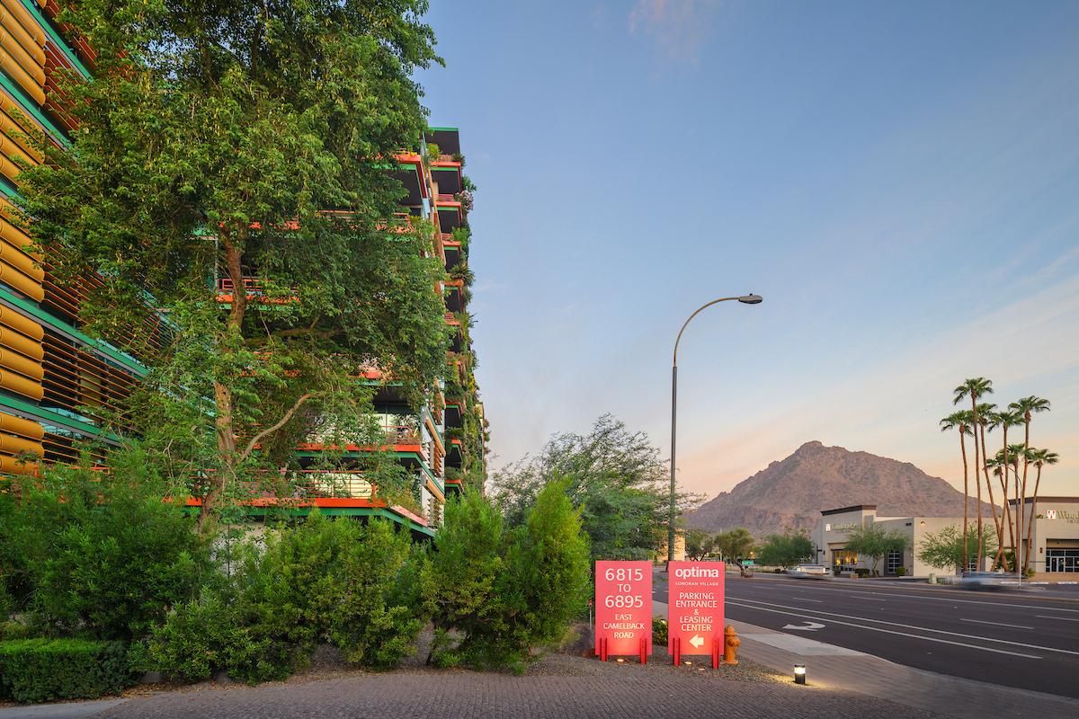 Modern building with lush greenery, street, red signs, and mountain views—Optima Sonoran Village embraces biophilic design.