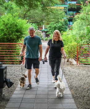 Man and woman walking two small dogs on a path, enjoying urban living amid greenery and red railings.