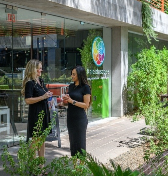 Two women stand and talk, holding drinks, outside a building with a colorful "Kaleidoscope" sign—a hub of urban living.