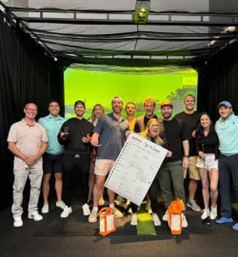 A group of people pose indoors with a large score sheet and gift bags, celebrating at an Optima Communities event.