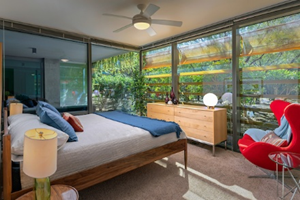 Modern bedroom with large windows, a wooden bed, dresser, red chair, and plants outside.