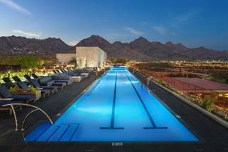 Rooftop swimming pool at dusk with lounge chairs, city lights, and mountains in the background.