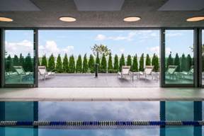 Indoor pool with lane divider, open doors lead to outdoor patio with lounge chairs and green hedges.