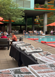 Pizza boxes on tables near a pool at Optima Communities, with people lounging under orange umbrellas during a lively community event.
