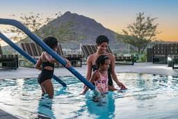 A woman and two children enjoy a swimming pool at sunset, with a mountain and lounge chairs in the background.