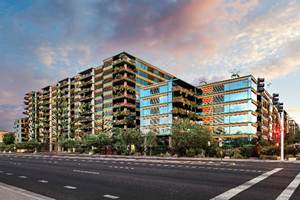 Modern apartment building with glass balconies and greenery, set against a colorful sky along a wide, empty street.
