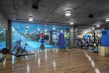 Modern gym with wooden floors, exercise bikes, rowing machines, and a glass wall overlooking an indoor swimming pool.