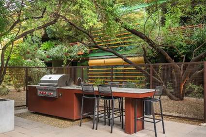 Outdoor kitchen with a grill, sink, and bar stools under leafy trees, surrounded by a fence and greenery.