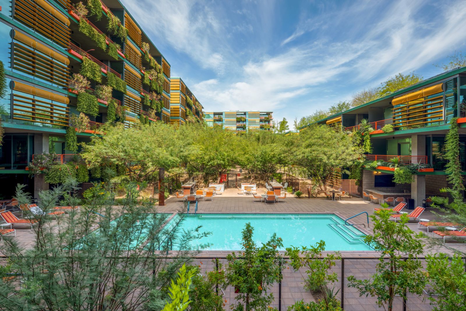 Modern apartment complex with green balconies and a central outdoor pool surrounded by trees and lounge chairs at Optima Sonoran Village apartments.