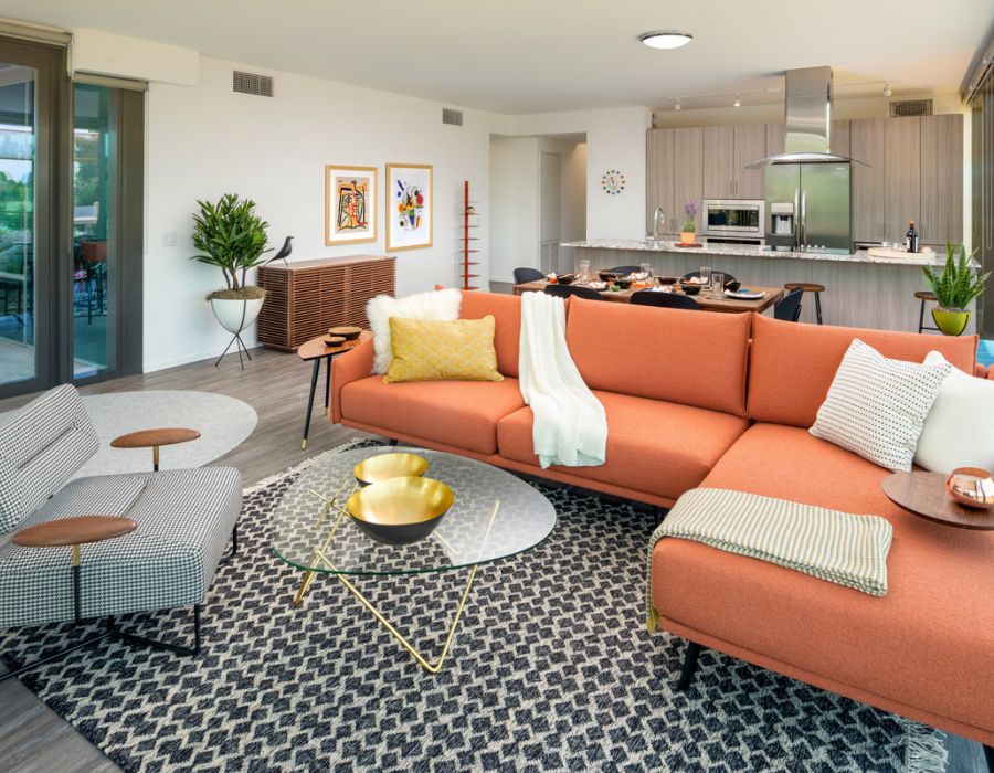 Modern living room with an orange sectional sofa, glass coffee table, and open kitchen in the background at Optima Sonoran Village apartments.