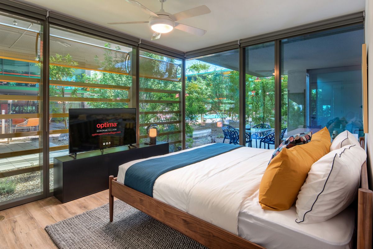 Optima Sonoran Village apartment Modern bedroom with large windows, a bed with yellow pillows, a wall-mounted TV, and a view of a garden patio.