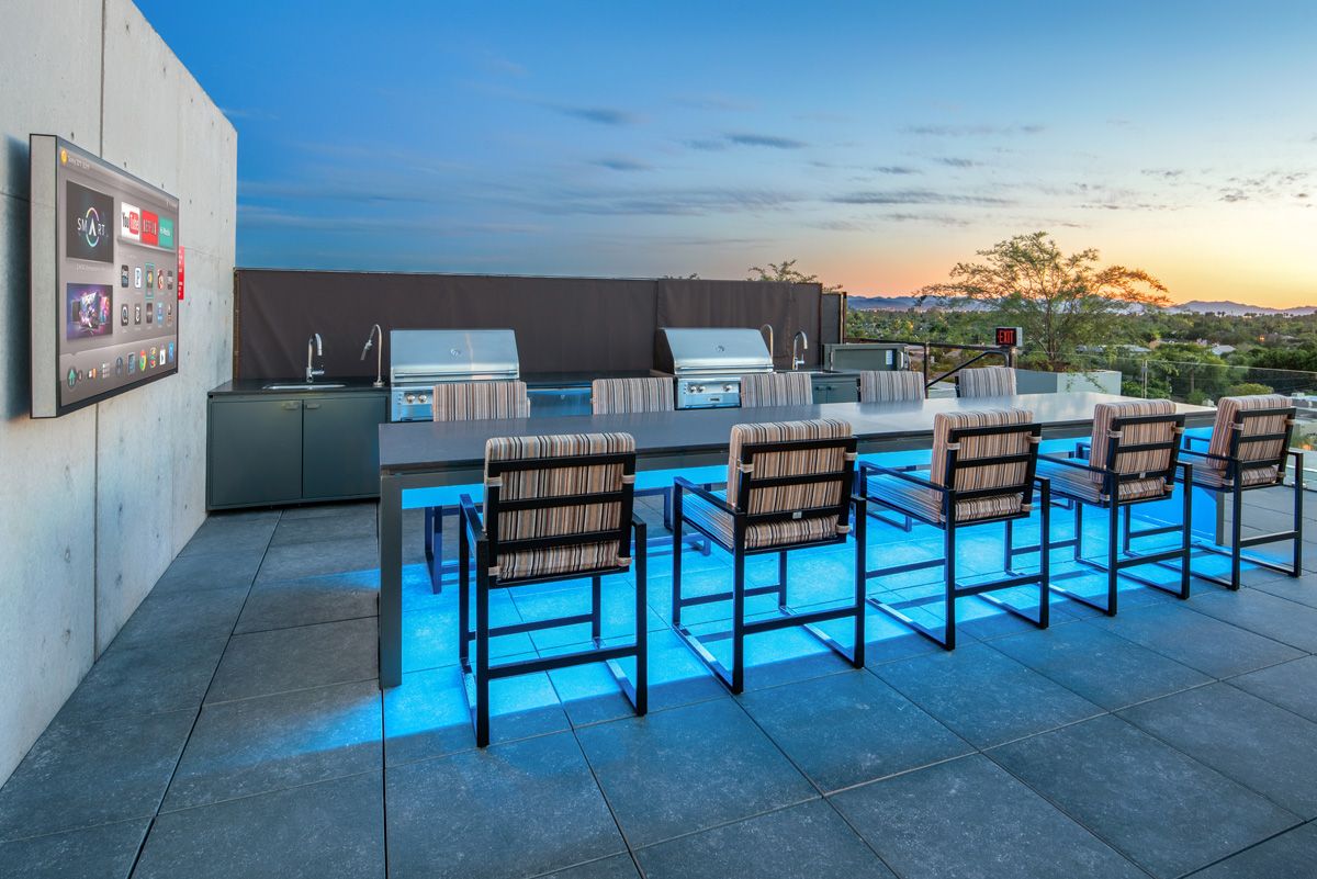 Modern outdoor kitchen with a long table, bar chairs, grills, and a TV, set against a sunset backdrop at Optima Sonoran Village apartments.