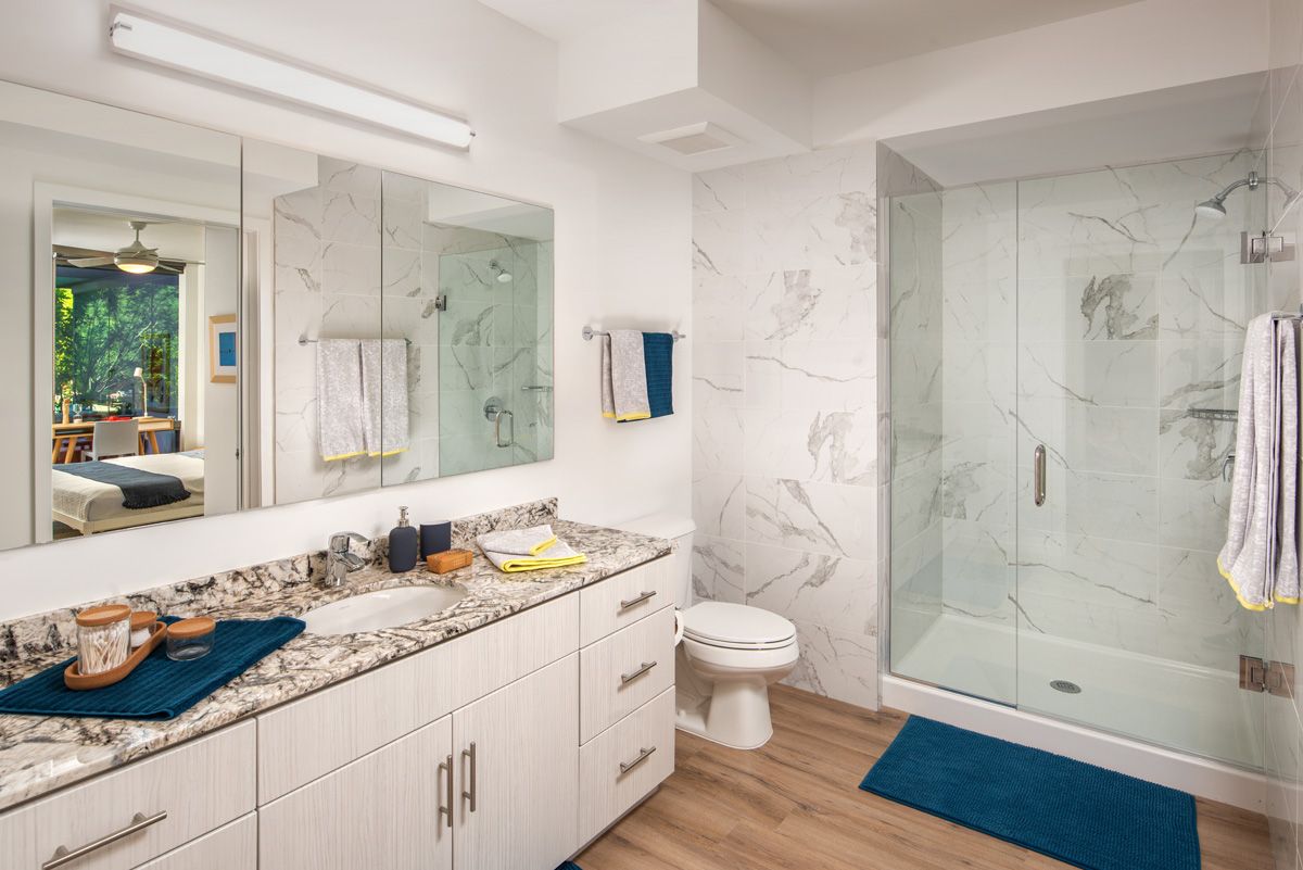 Optima Sonoran Village apartment Modern bathroom with marble countertops, glass shower, and wooden floor. Towels and decor in blue accents.