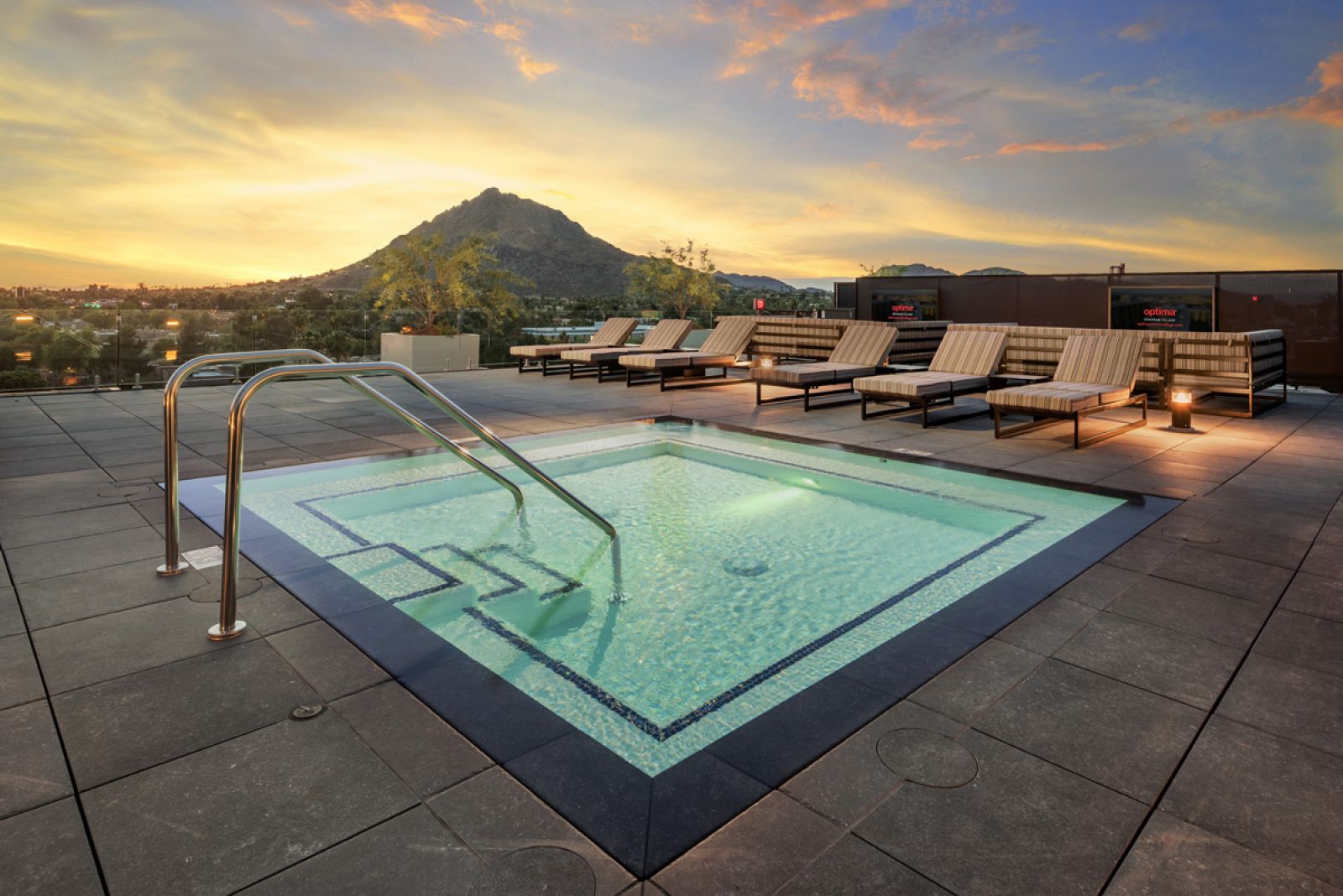 Rooftop pool with lounge chairs, overlooking a sunset-lit mountain and cityscape at Optima Sonoran Village apartments.