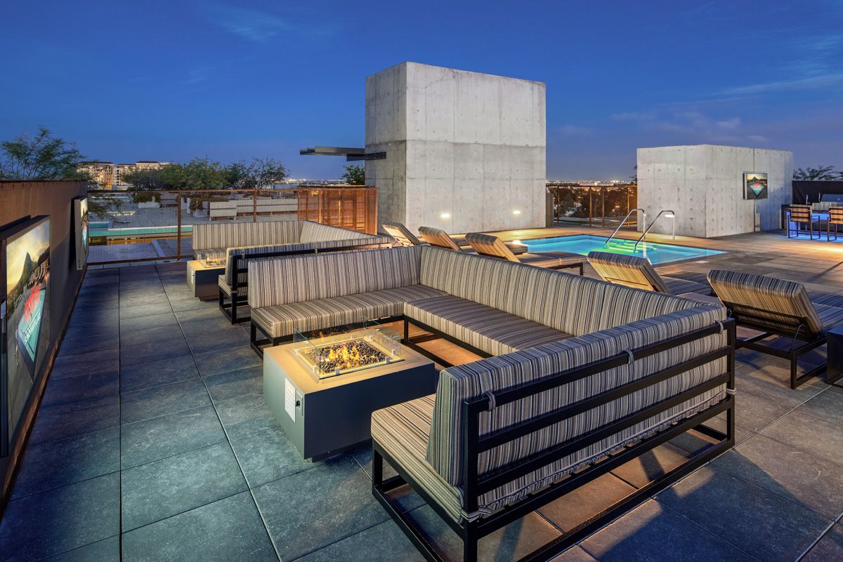 Rooftop patio with striped seating, fire pit, pool, and outdoor dining area against a twilight sky backdrop at Optima Sonoran Village apartments.