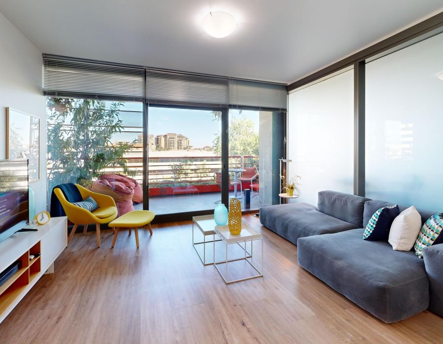 Optima Sonoran Village apartments modern living room with a gray sofa, TV, colorful chairs, and balcony with city view. Natural light fills the space.