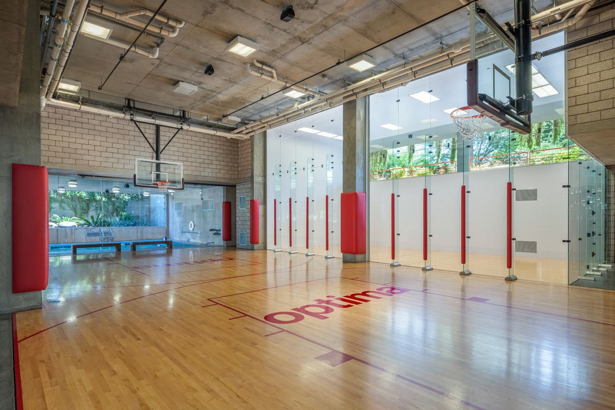 Indoor basketball court with glass walls, a wooden floor, nets, and padded red accents at Optima Sonoran Village apartments.